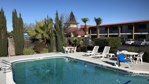A clear swimming pool with lounge chairs and a table, surrounded by tall trees and a building with a red roof. - Super 8 by Wyndham Corning (Corning)