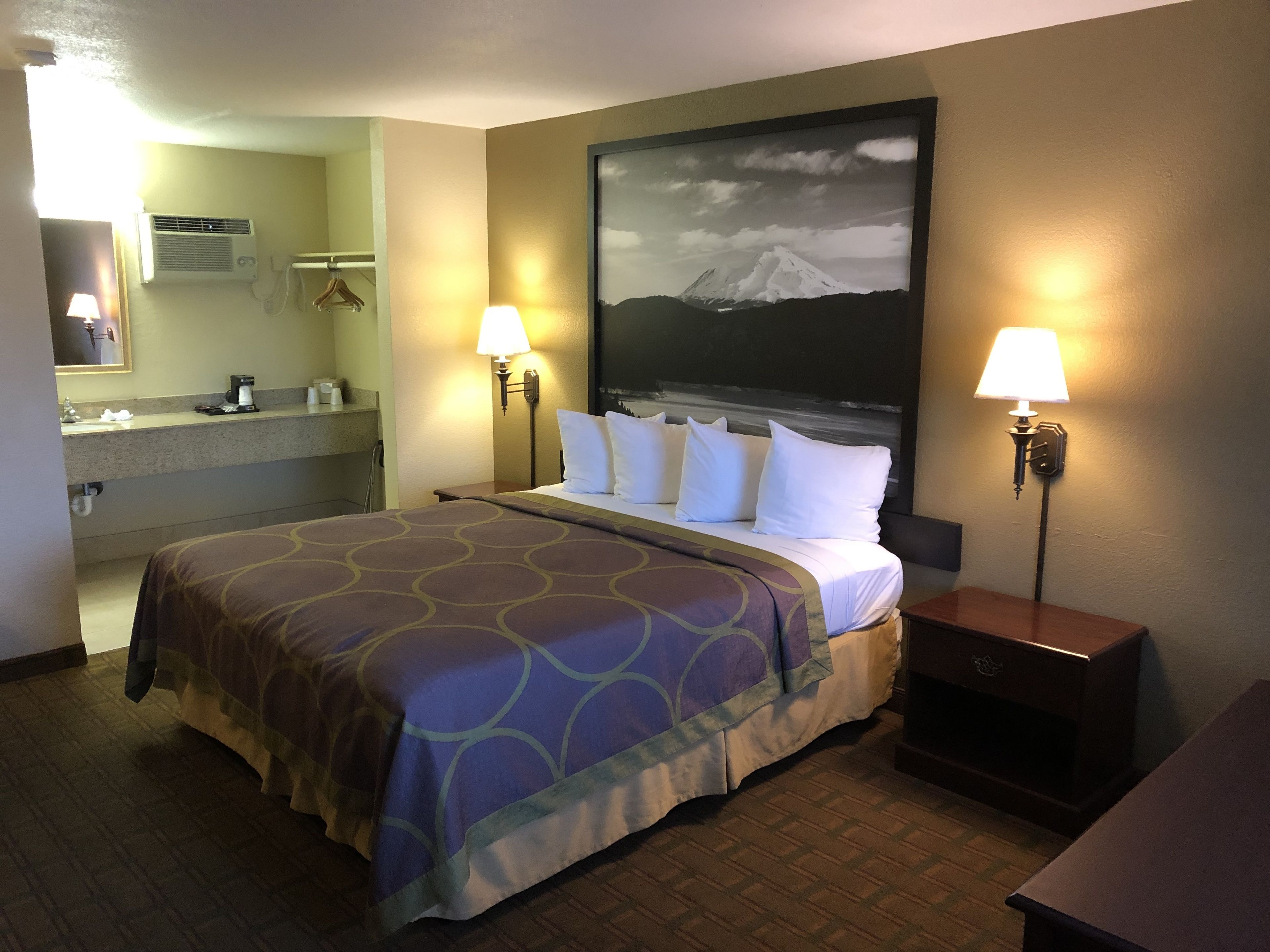 Een hotelkamer met een bed, twee bedlampen, een nachtkastje, een schilderij van een berg, een badkamer met een wastafel en spiegel, en een airconditioner.