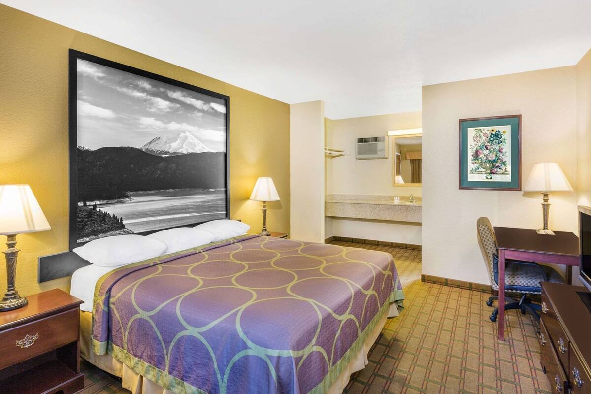 A hotel room with a bed, two bedside lamps, a desk with a chair, a bathroom with a sink and mirror, and a large framed picture on the wall.
