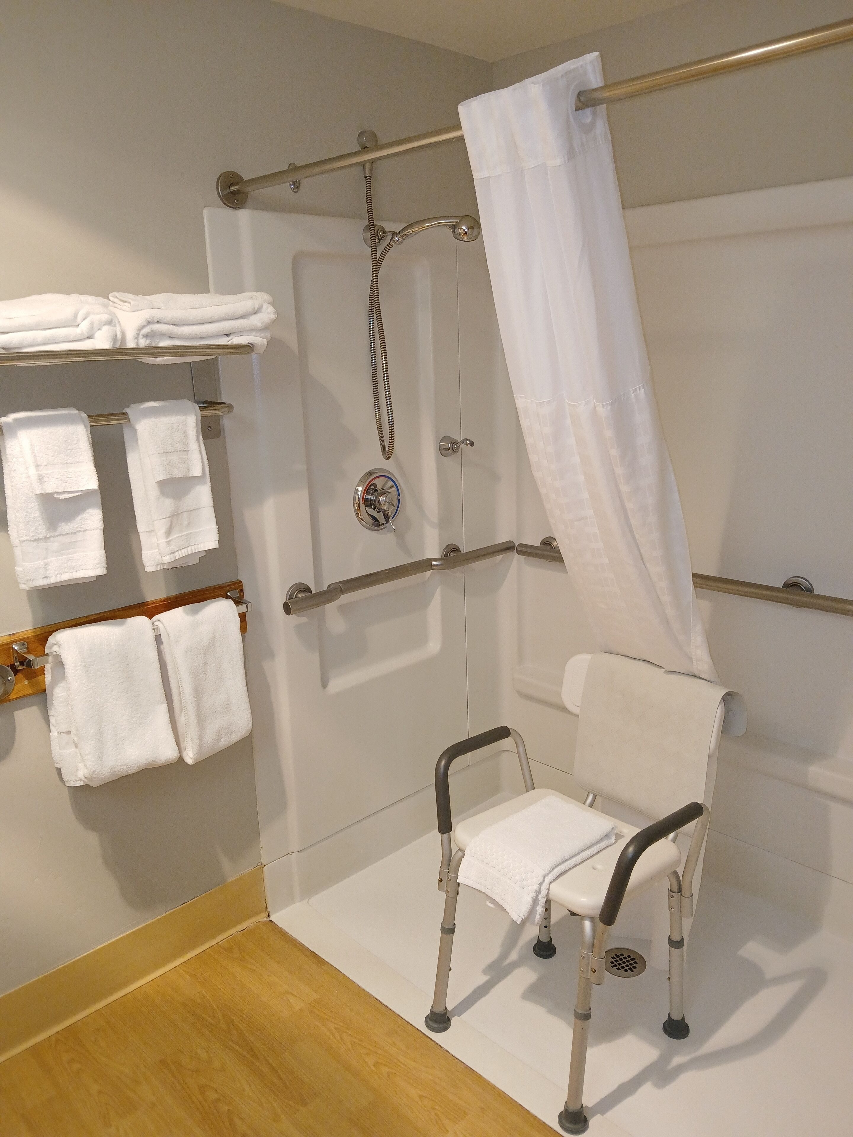 A shower area with a grab bar, a white shower curtain, and a white chair.