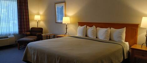 Habitación de hotel con una cama grande, dos mesitas de noche con lámparas, una silla, una pequeña mesa y una ventana con cortinas.