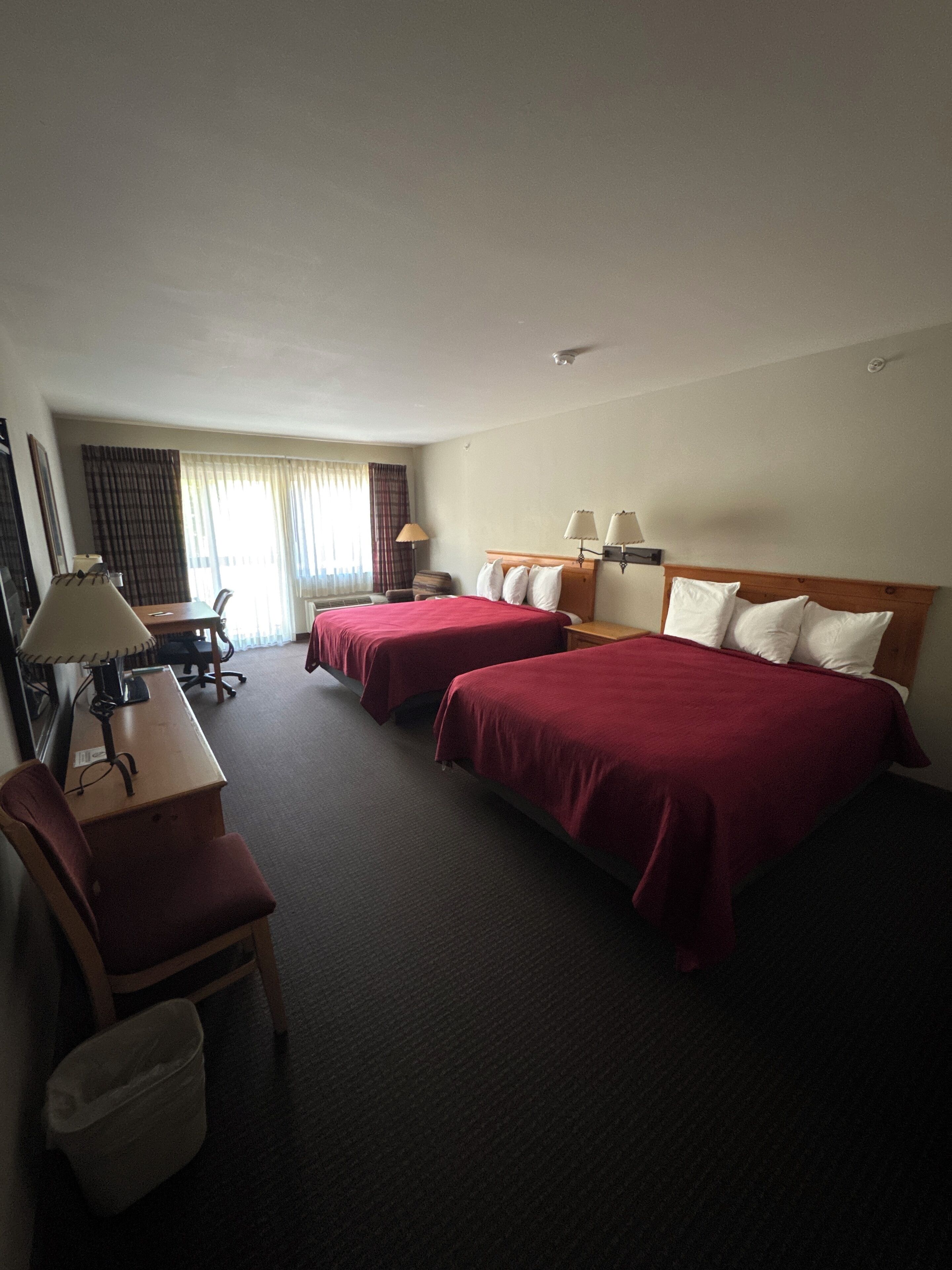 Une chambre d’hôtel avec deux lits, un bureau, une chaise, une lampe et une fenêtre avec des rideaux.