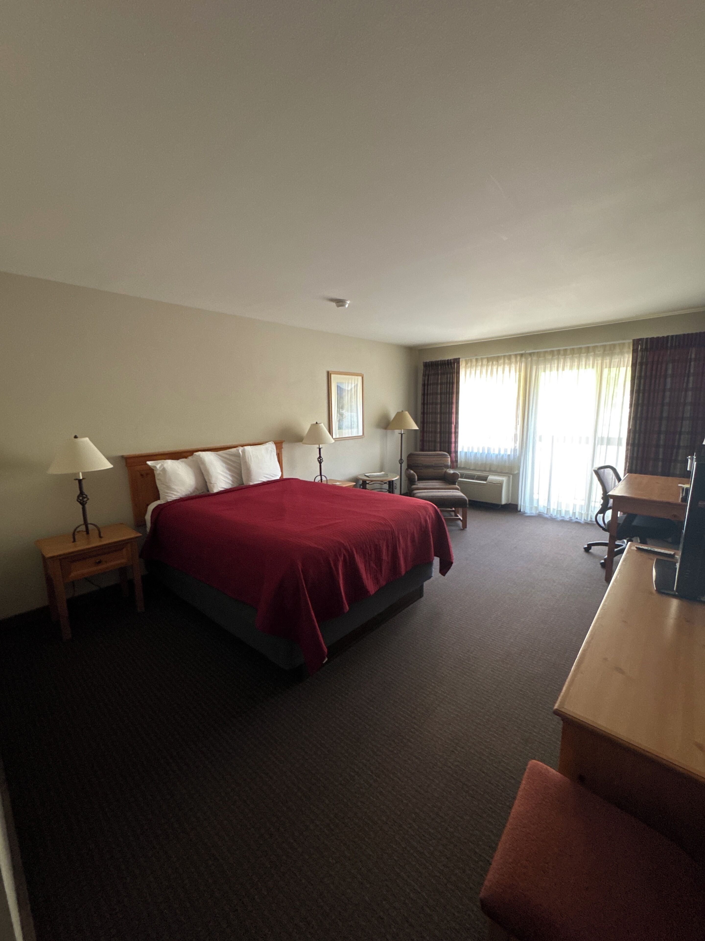A hotel room with a bed, two bedside tables with lamps, a desk, a chair, and a window with curtains.