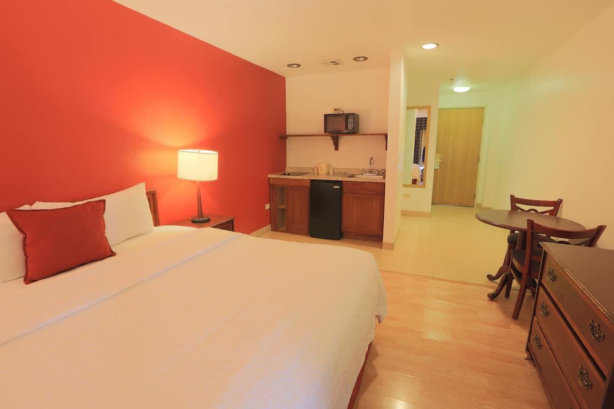 A hotel room with a bed, a desk, a chair, a small kitchenette, and a door leading to another room.
