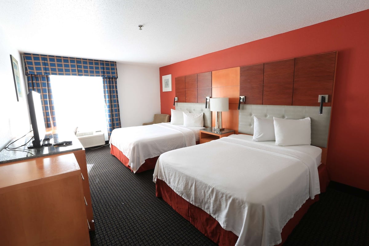 A hotel room with two beds, a desk, a TV, and a window with blue curtains.