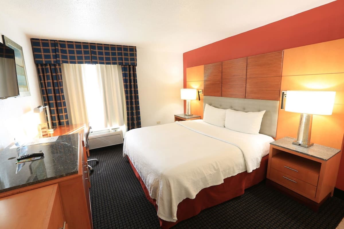 A hotel room with a bed, bedside tables, a desk, a chair, a TV, and a window with curtains.