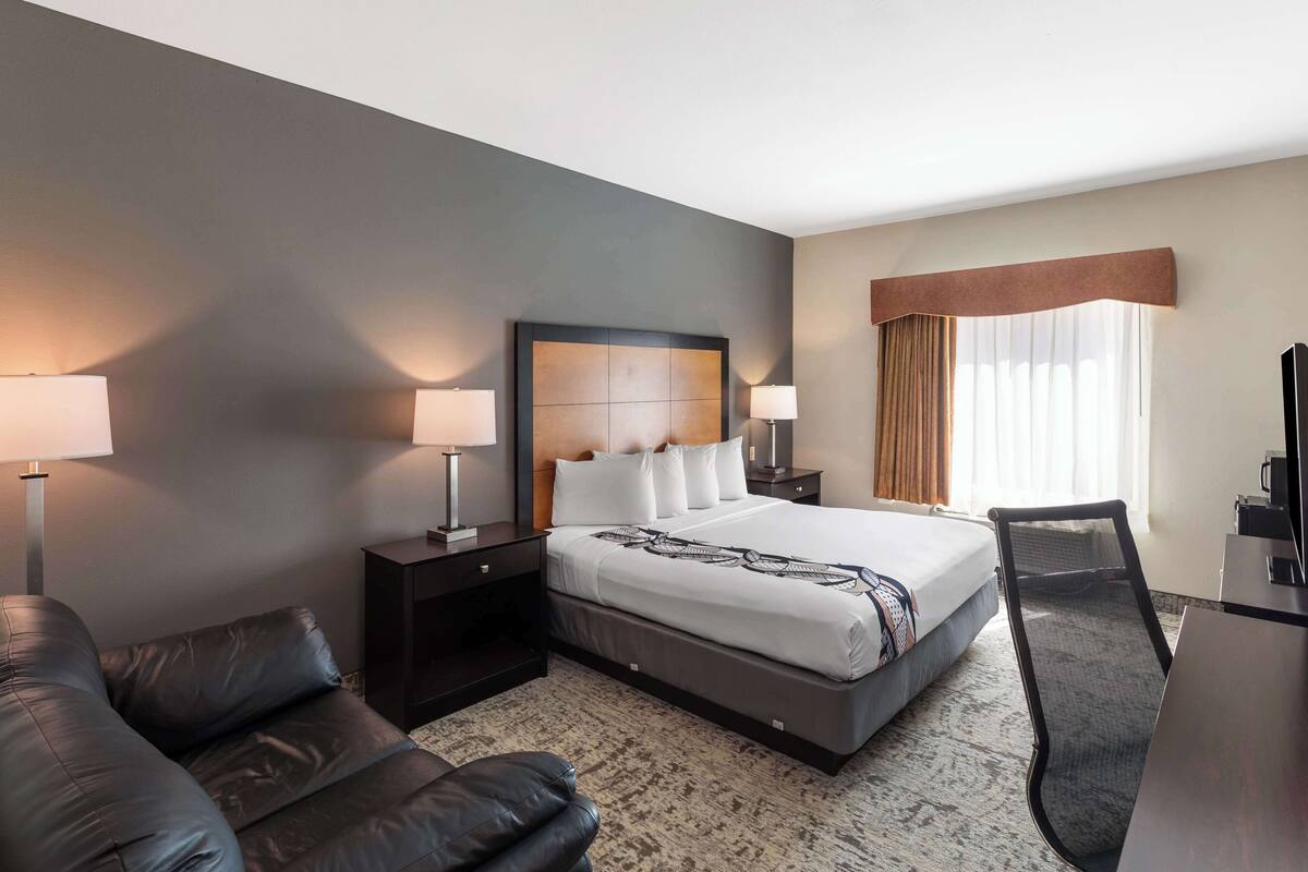 A modern hotel room with a large bed, two bedside tables with lamps, a leather sofa, a desk, and a chair.