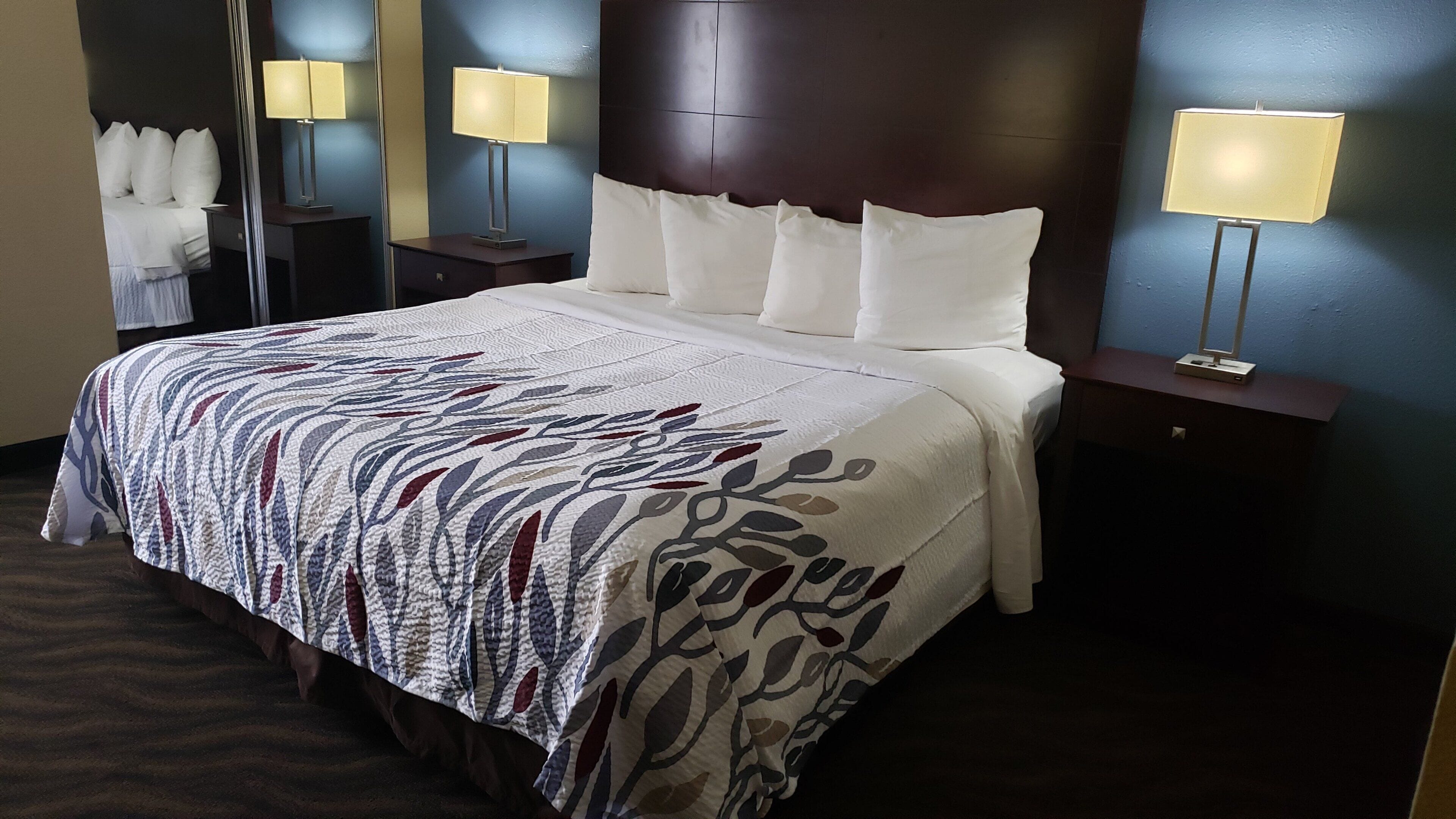Una habitación de hotel con una cama grande, cabecera, dos mesitas de noche con lámparas y un espejo.