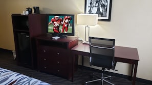 A hotel room with a television on a wooden cabinet, a desk with a chair, a lamp, and a bed with a patterned bedspread. - Red Roof Inn Port Allen West LSU Stadium (Port Allen)