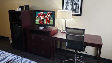 Habitación de hotel con una televisión sobre un mueble de madera, un escritorio con una silla, una lámpara y una cama con colcha estampada.