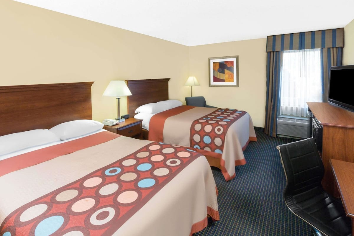 Quarto de hotel com duas camas, uma escrivaninha com televisão, uma luminária e um quadro na parede.