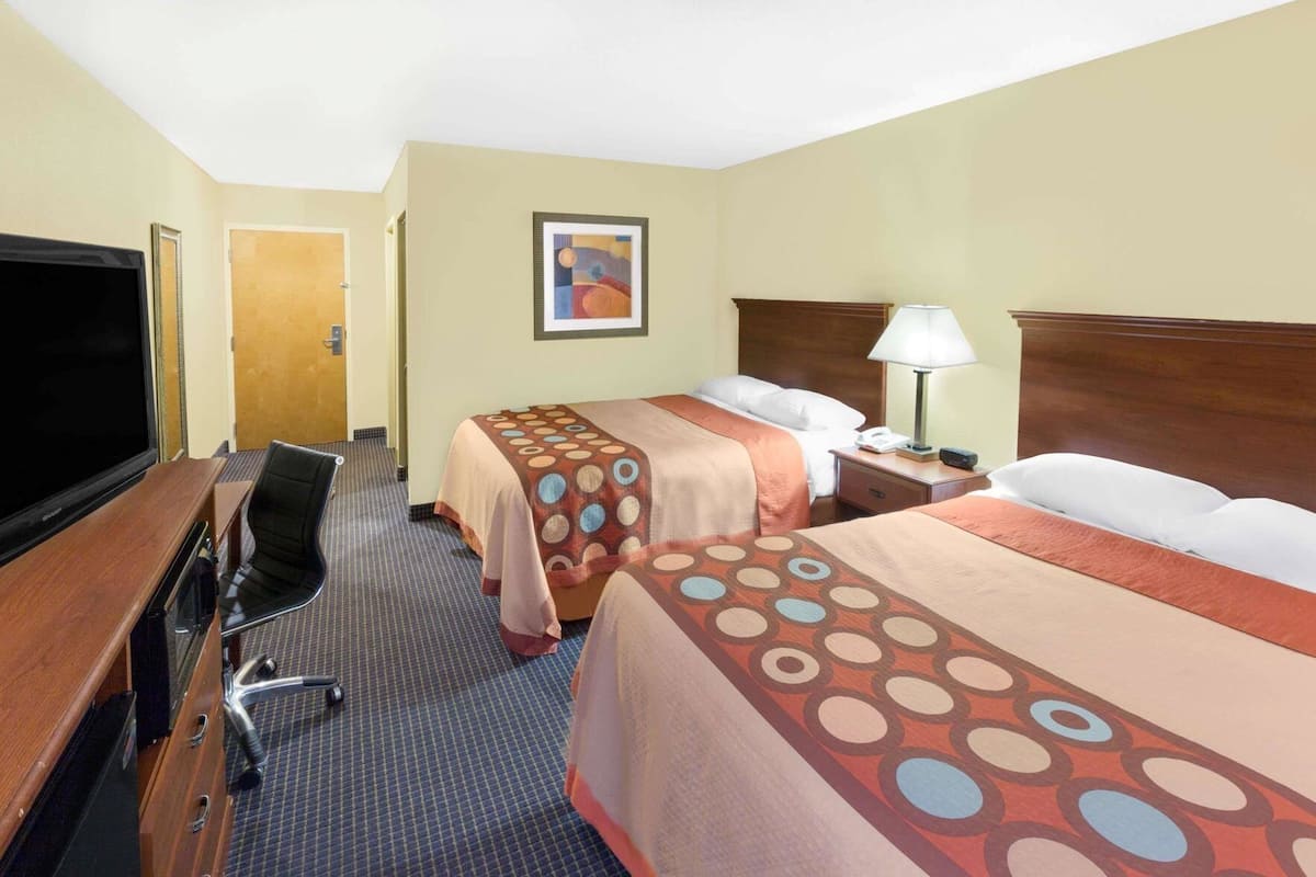 Quarto de hotel com duas camas, uma mesa de trabalho com televisão, uma cadeira, um abajur e um quadro na parede.