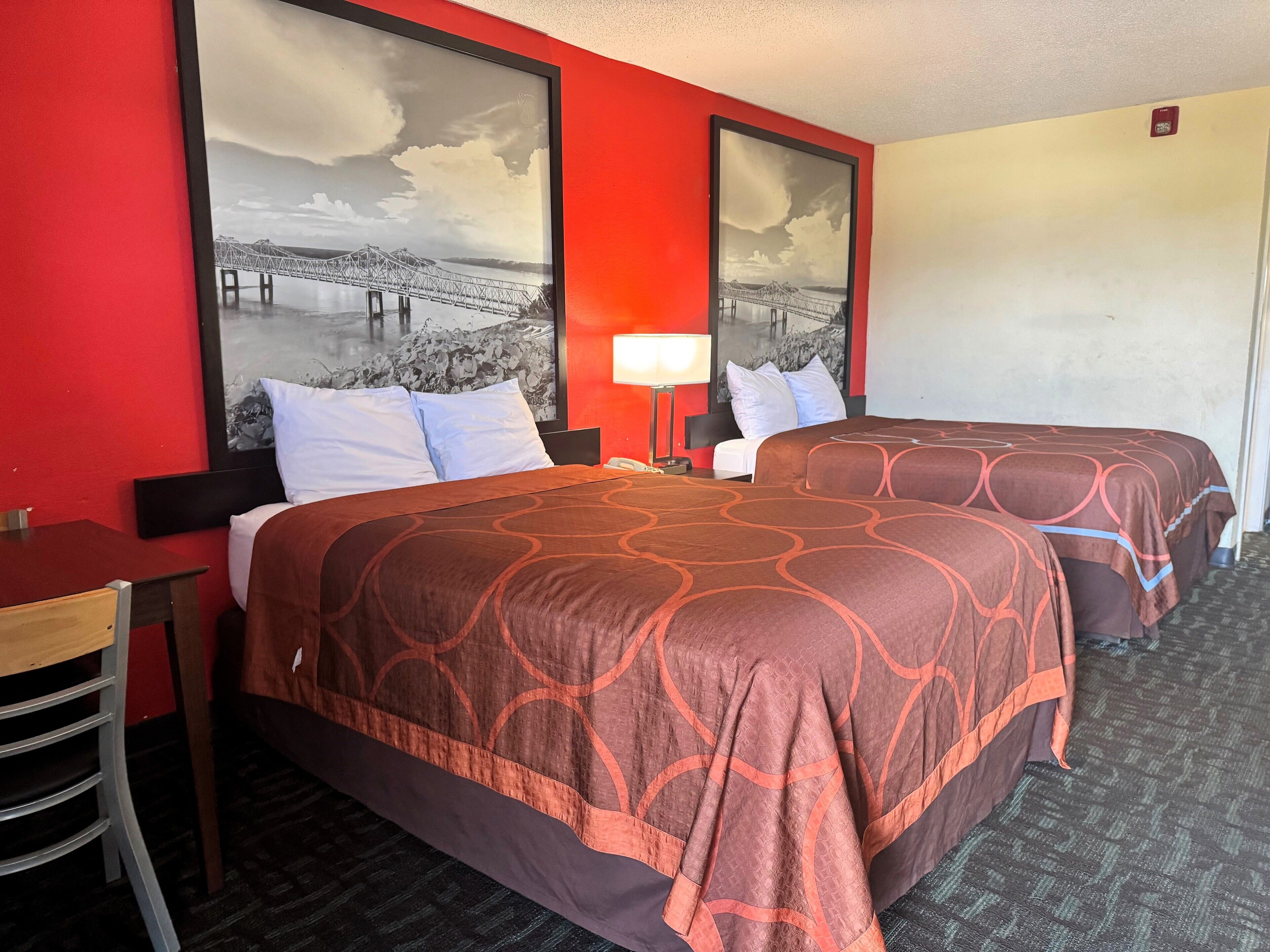 A hotel room with two beds, a desk, and a chair. There are two large framed pictures on the wall, one of a bridge and one of a mountain range.