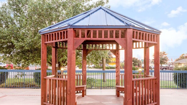 A wooden gazebo with a metal roof, surrounded by a blue fence and trees.