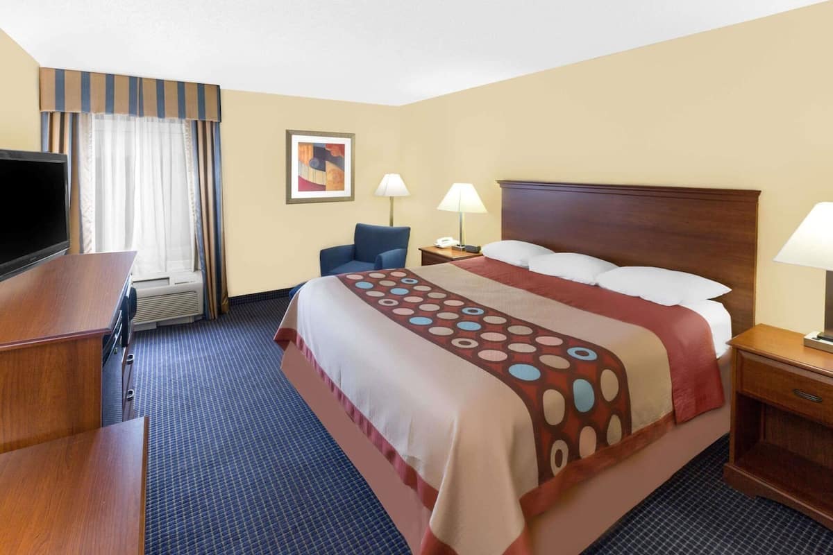 Quarto de hotel com uma cama, duas luminárias laterais, uma escrivaninha com televisão, uma cadeira e uma janela com cortinas.