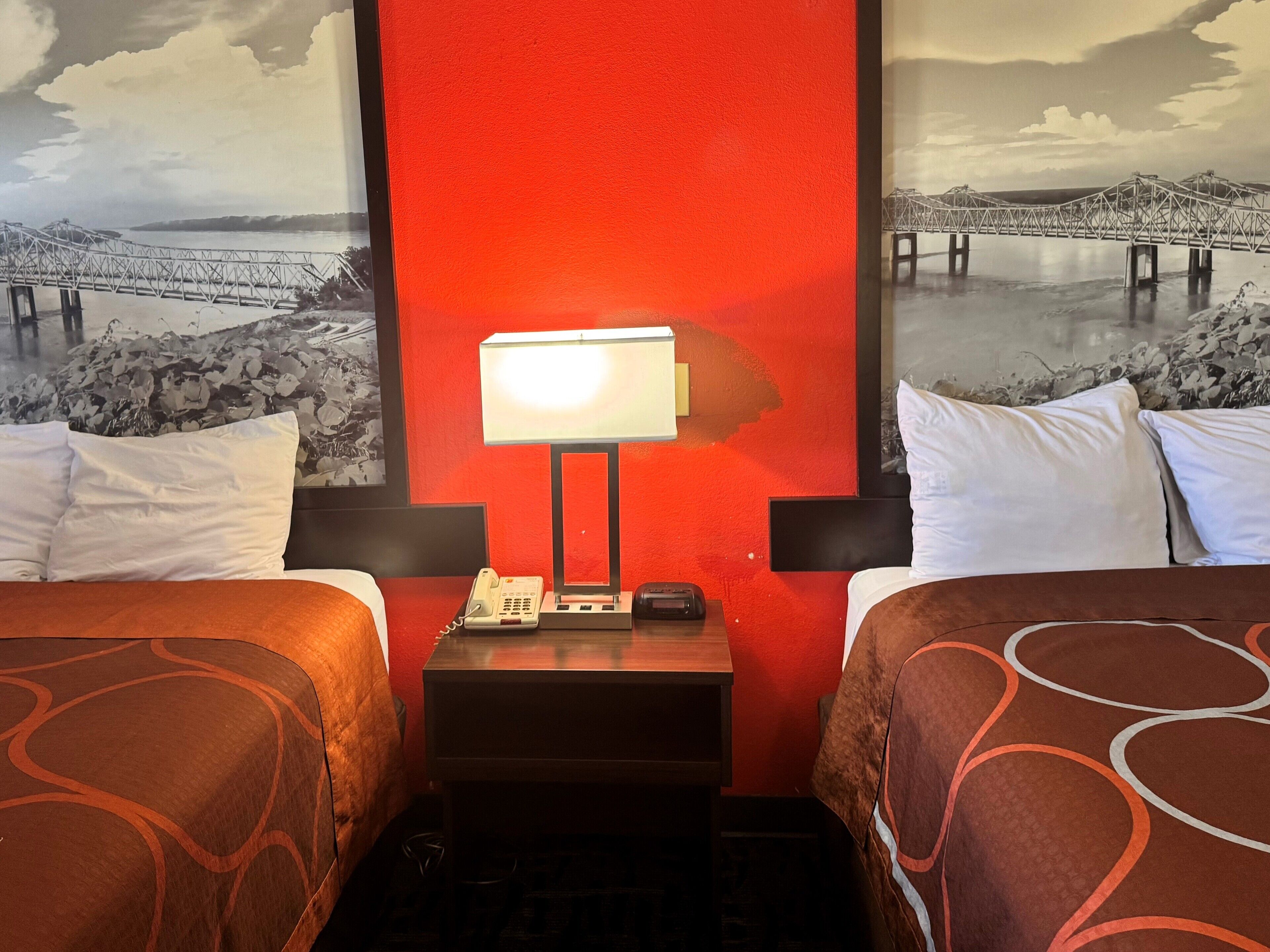 Ein Hotelzimmer mit zwei Betten, einem Nachttisch mit Lampe, einem Telefon und einem Wandbild, das eine Brücke zeigt.