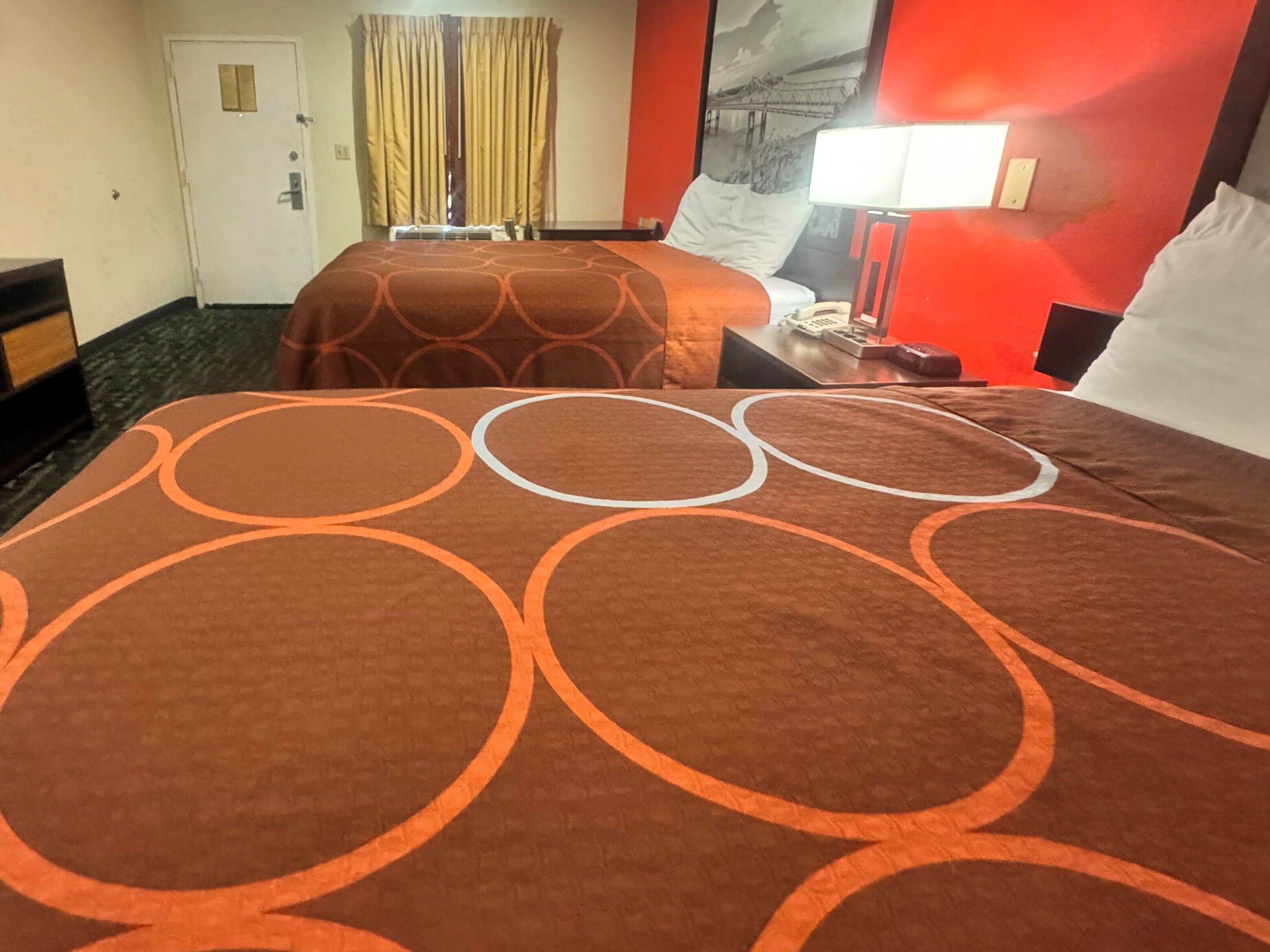 A hotel room with two beds, a red accent wall, a window with curtains, a bedside table with a lamp, and a door with a handle.