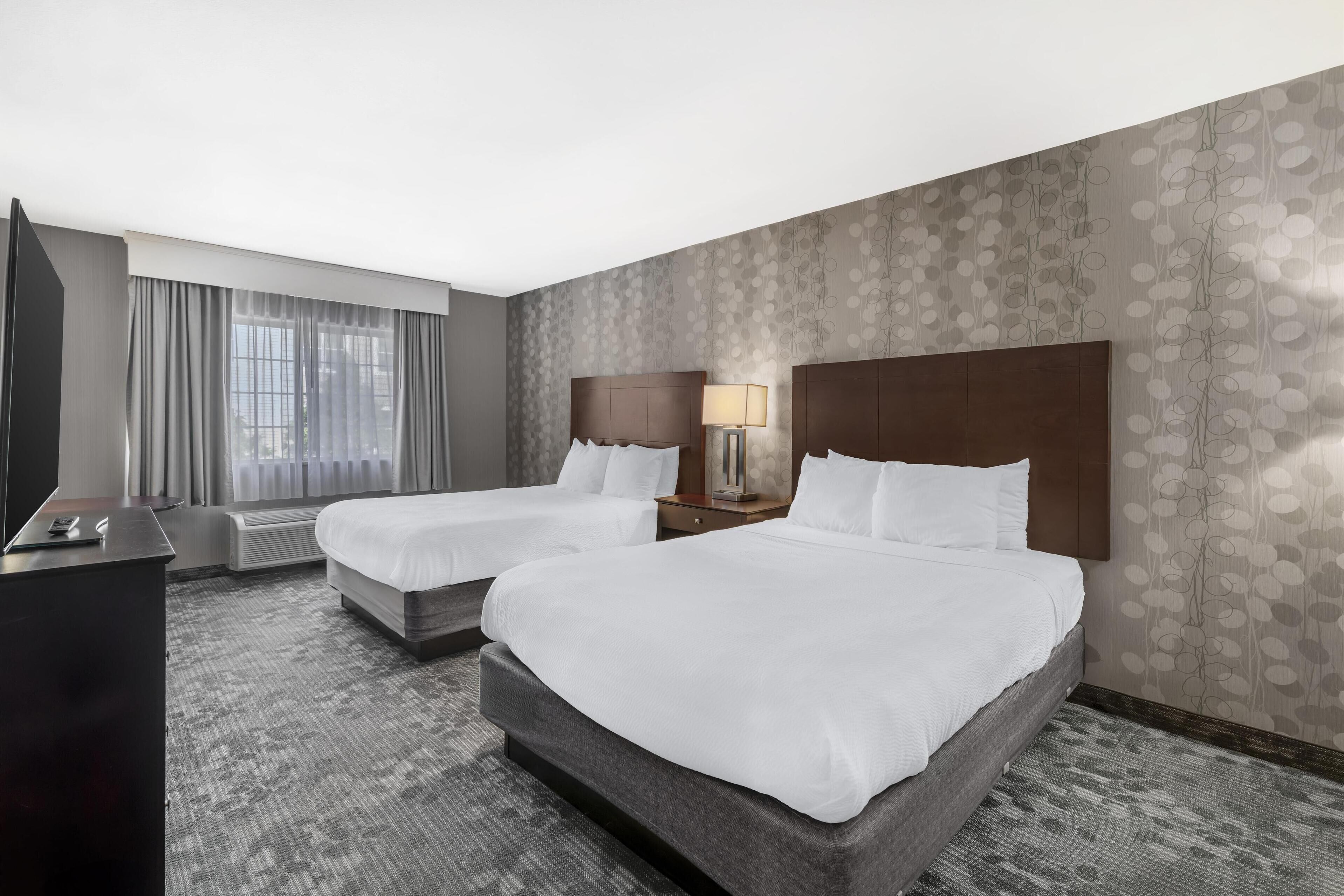 A hotel room with two beds, a desk, a chair, and a large window with curtains.