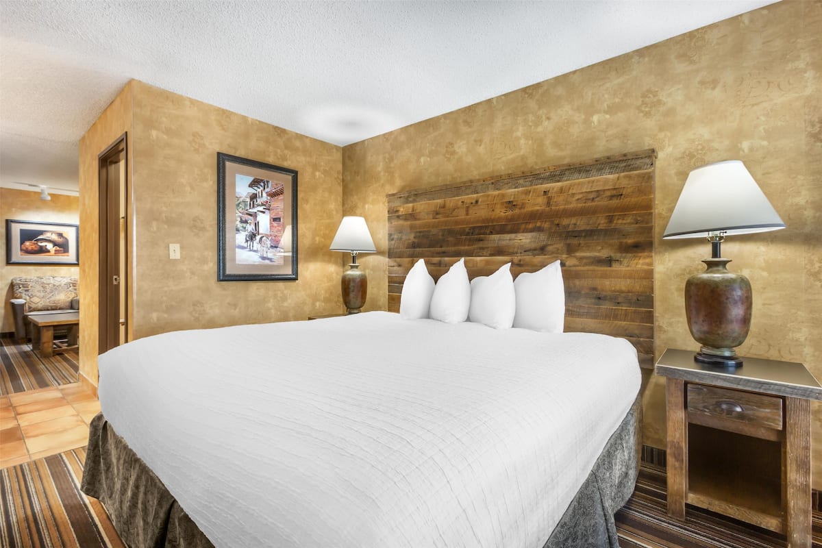 Une chambre d’hôtel avec un grand lit, deux lampes de chevet, une tête de lit en bois, un tableau encadré au mur et une petite table avec une chaise.