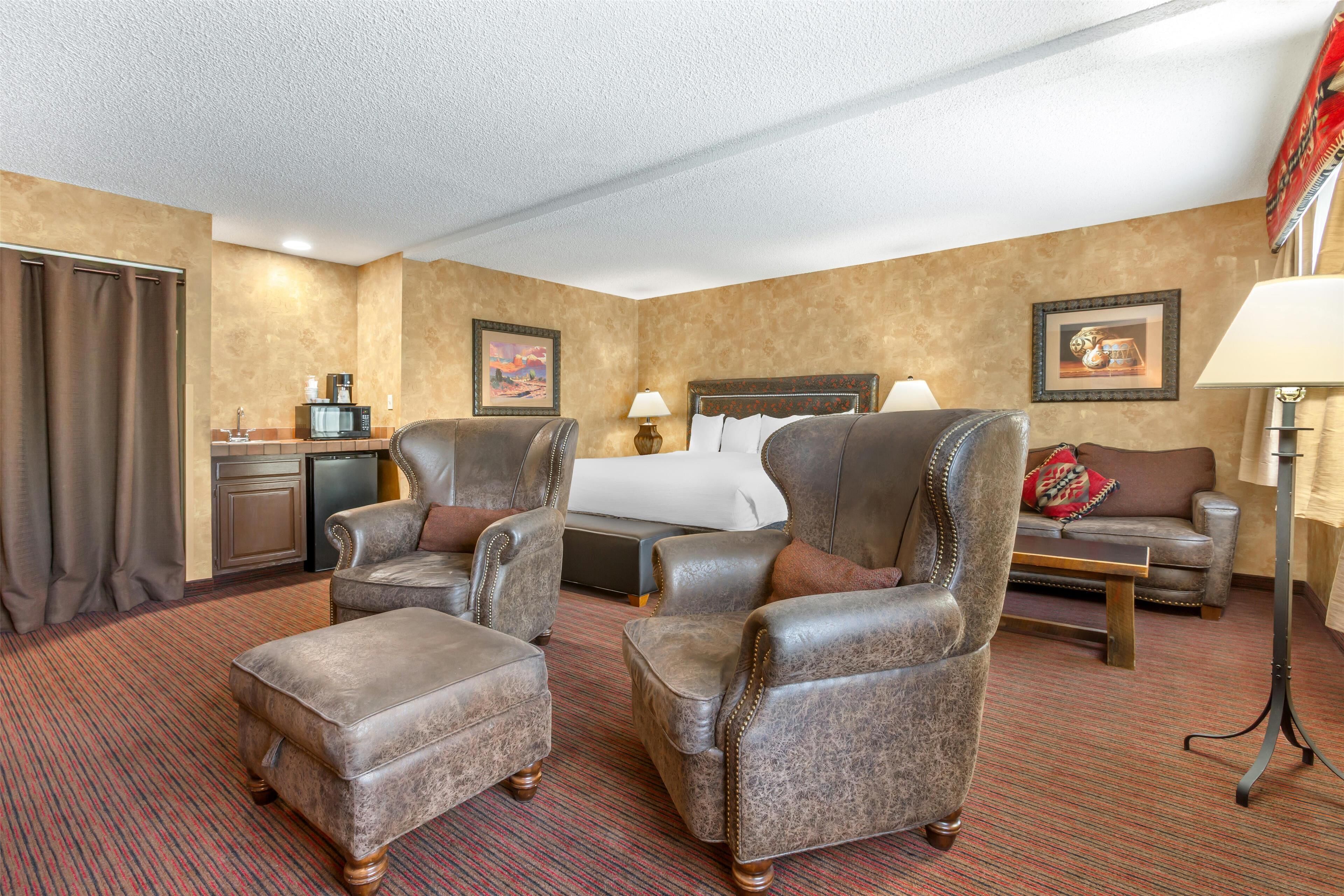 Une chambre d’hôtel avec un grand lit, deux fauteuils en cuir, une petite table, un canapé, un bureau avec une lampe et une machine à café.