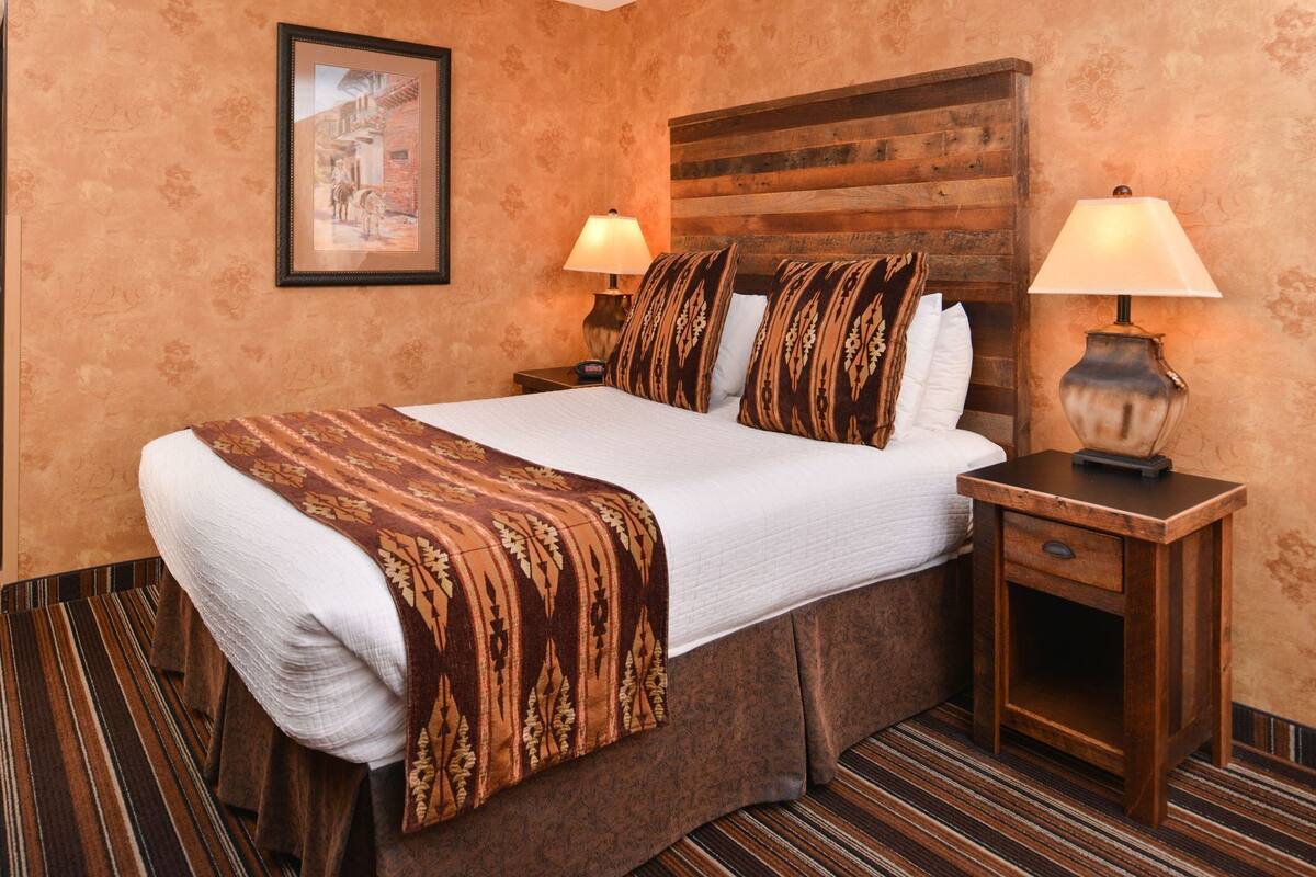 Une chambre d’hôtel avec un grand lit, deux tables de chevet, une tête de lit en bois, un tableau encadré au mur et un plaid à motifs.