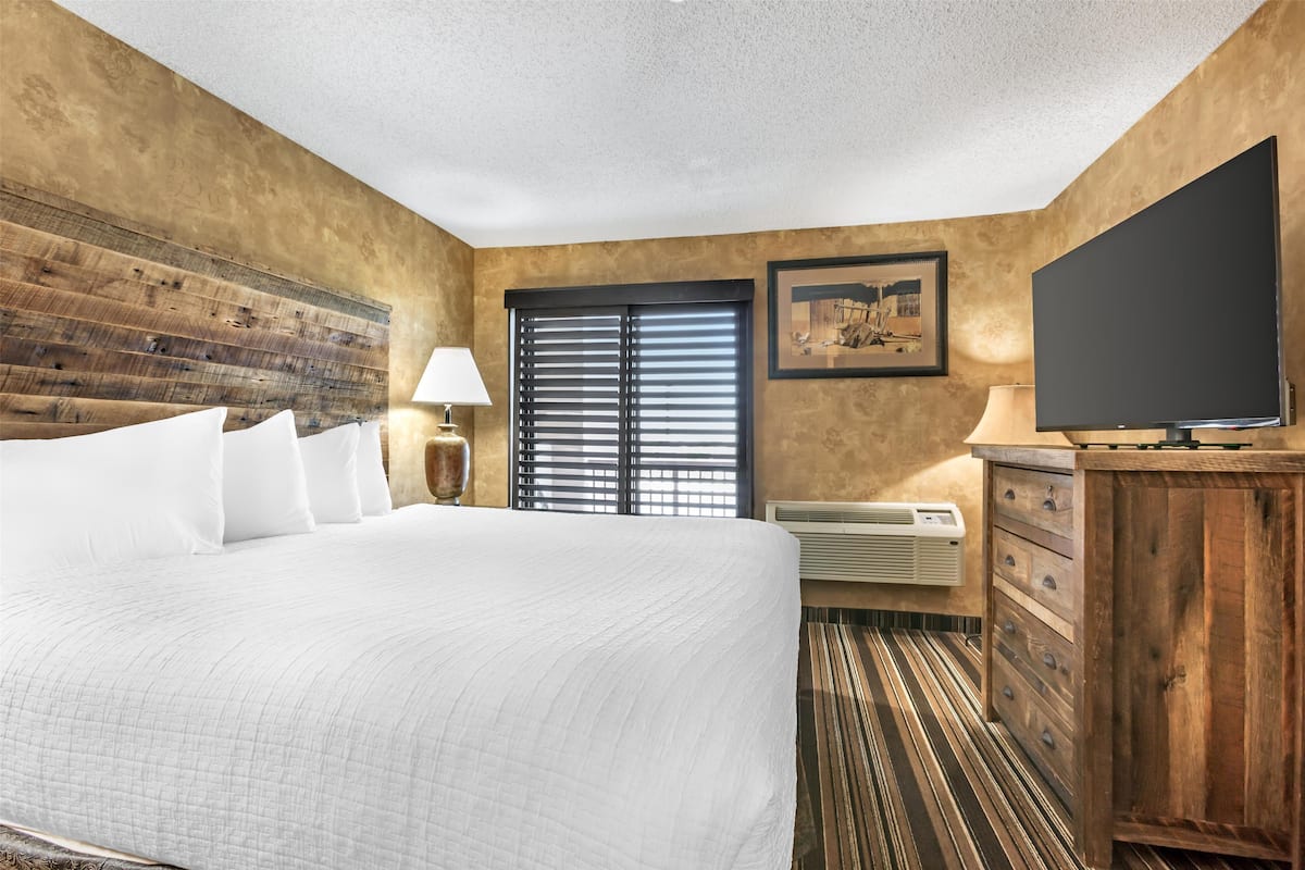 Une chambre d’hôtel avec un grand lit, une tête de lit en bois, un téléviseur à écran plat, une commode et une fenêtre avec des stores.