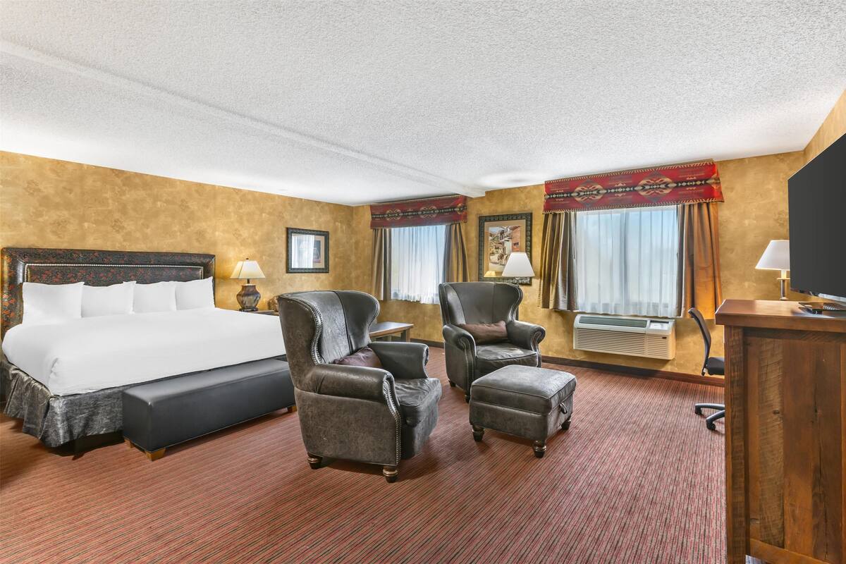 A hotel room with a large bed, two armchairs, a small table, and a desk with a computer.