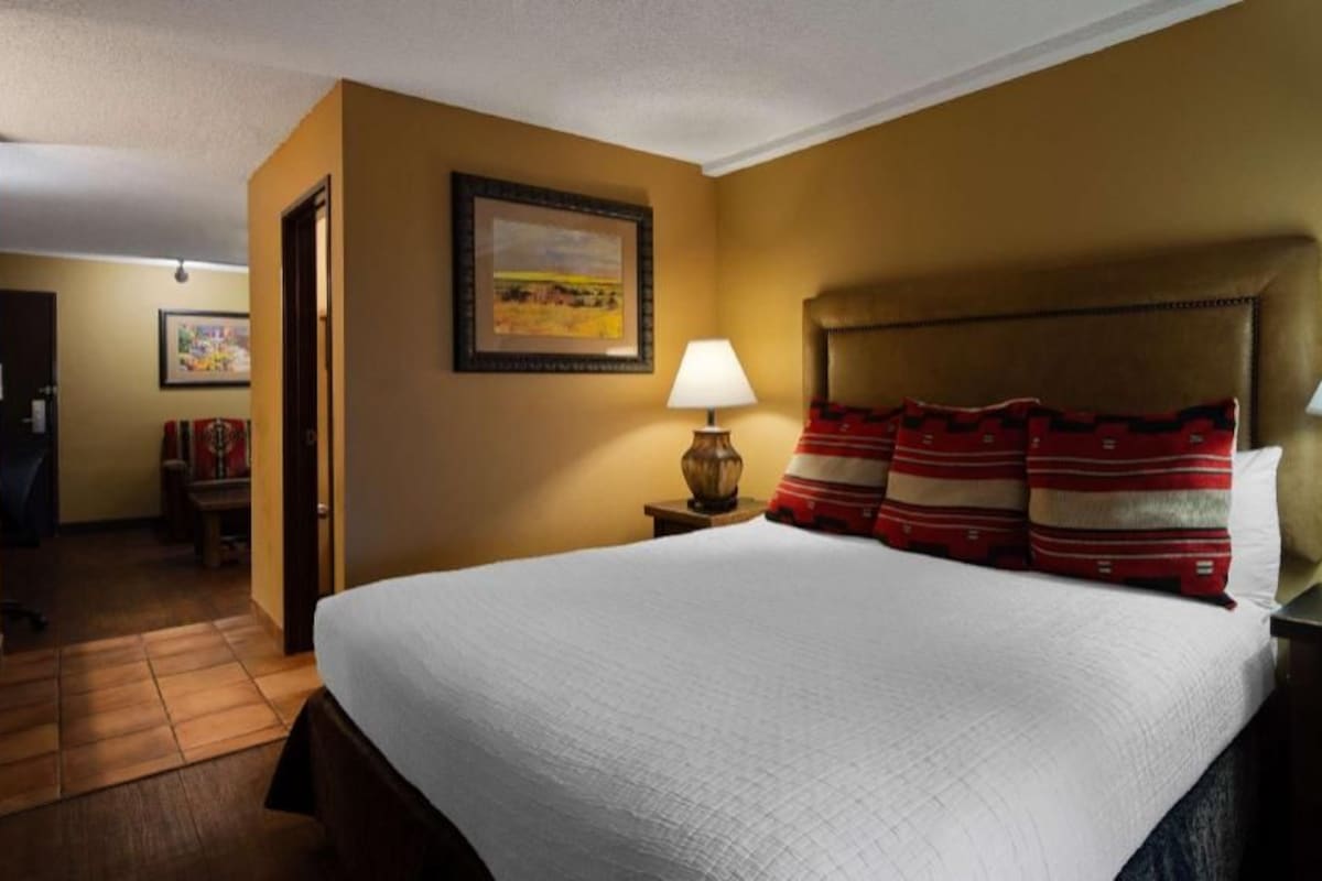 A hotel room with a large bed, a nightstand with a lamp, a framed picture on the wall, and a door leading to another room.