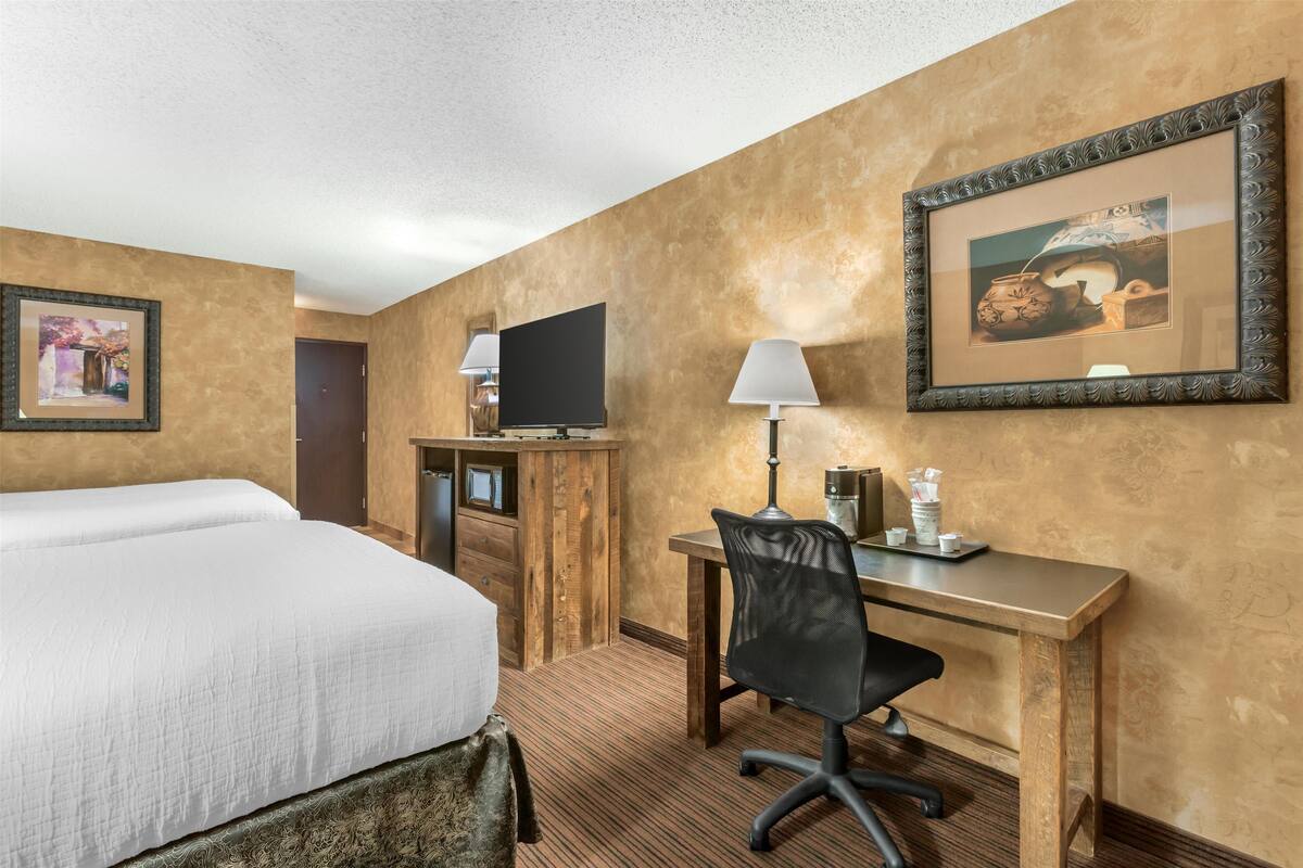 A hotel room with a large bed, a desk with a chair, a television, and framed artwork on the walls.