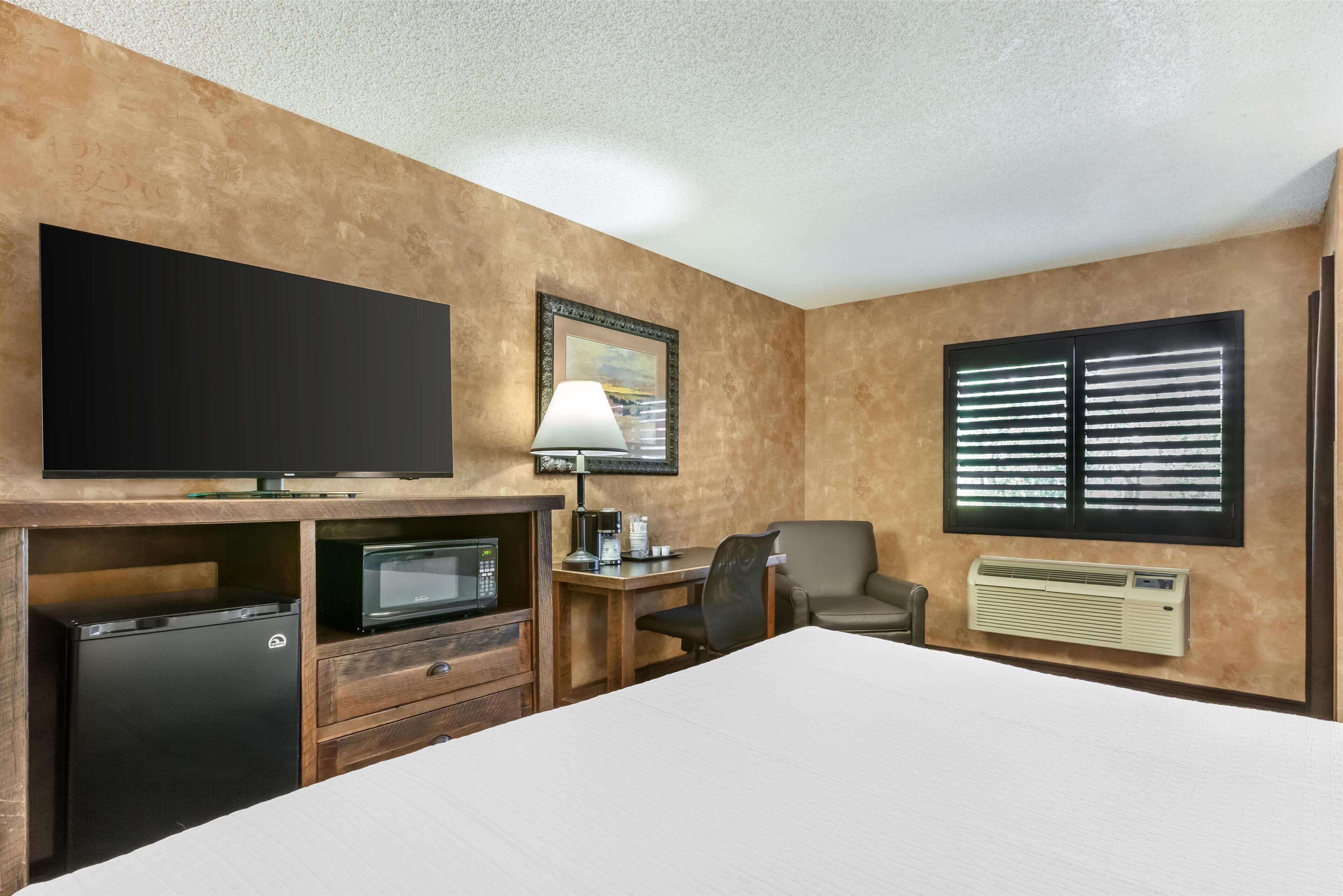 Une chambre d’hôtel comprenant un lit, un bureau avec une lampe, une télévision, un four à micro-ondes, un réfrigérateur, une chaise et une fenêtre avec des stores.