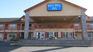 Un edificio de dos pisos con un cartel que dice “Rice Hill Inn”. Cuenta con fachada de ladrillo, un balcón con barandillas rojas y un techo plano.