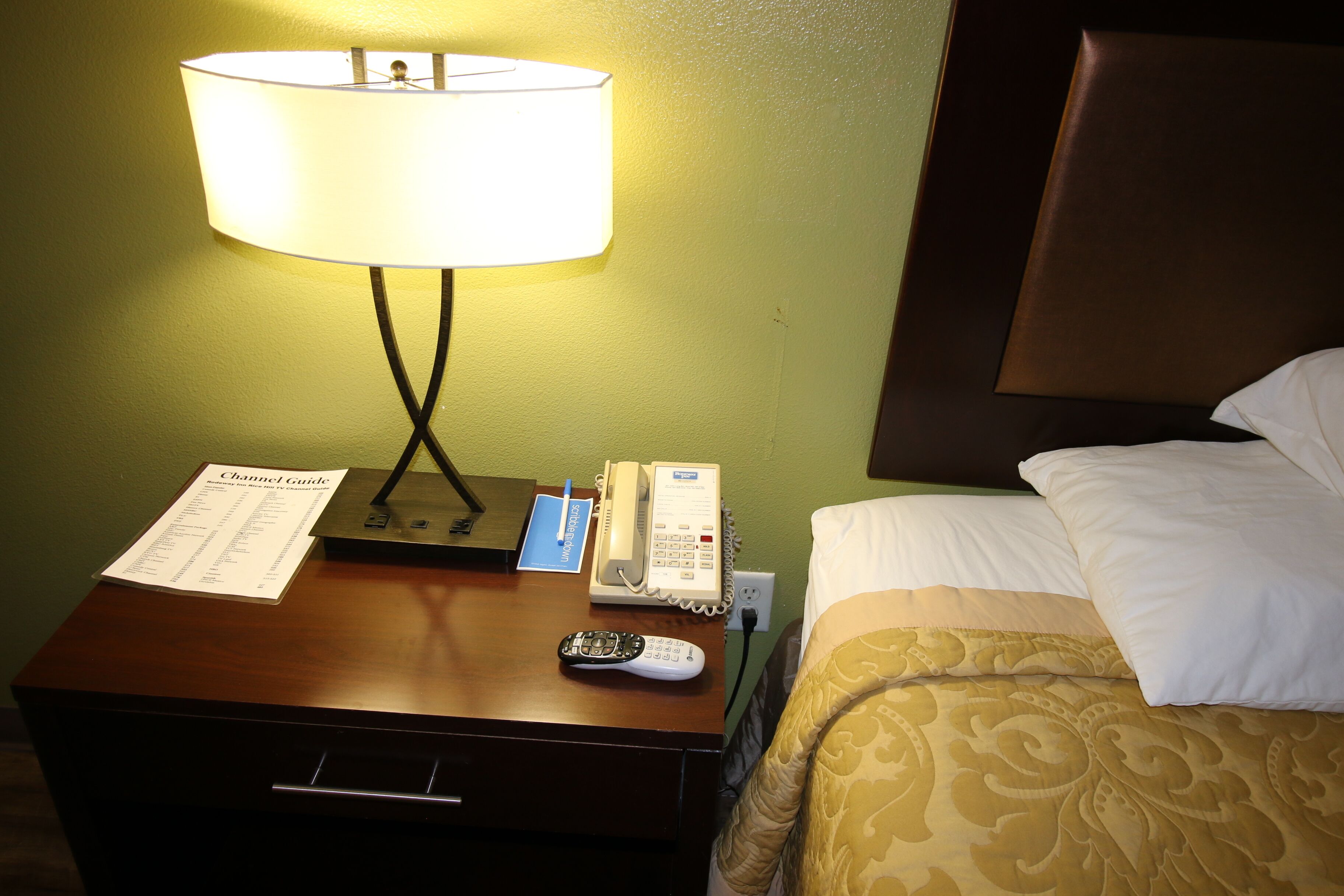 A hotel room with a wooden nightstand, a lamp, a remote control, a telephone, and a bed with a patterned bedspread.