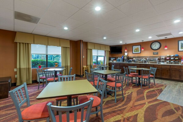 Une salle à manger d’hôtel avec des tables et des chaises, un coin cuisine avec une horloge, et une vue par la fenêtre.