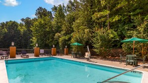 Une piscine rectangulaire avec un escalier en métal, entourée de chaises longues et de tables, et un espace boisé en arrière-plan.