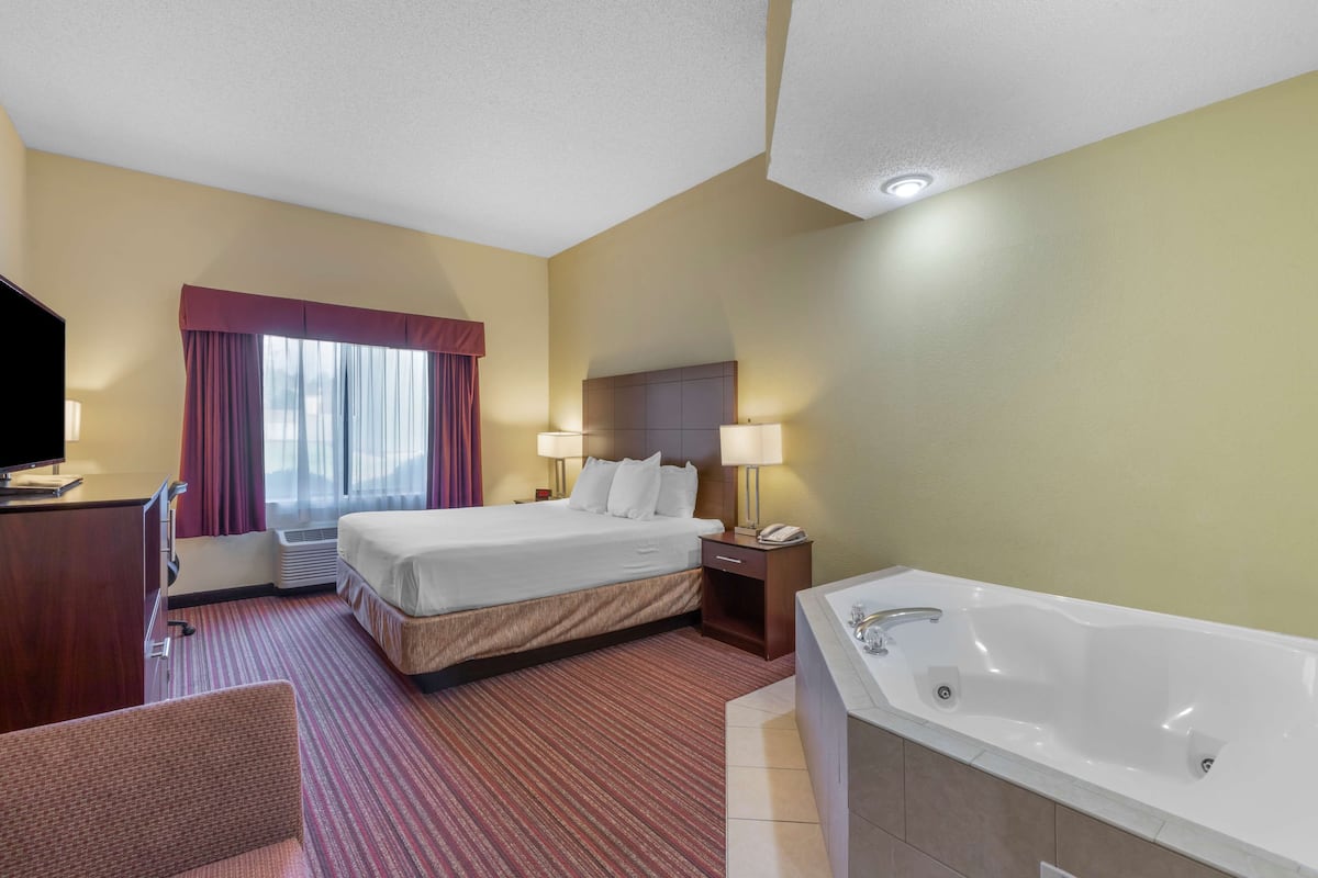 A hotel room with a bed, a desk, a chair, a large window with curtains, a bathroom with a bathtub, and a flat-screen TV.