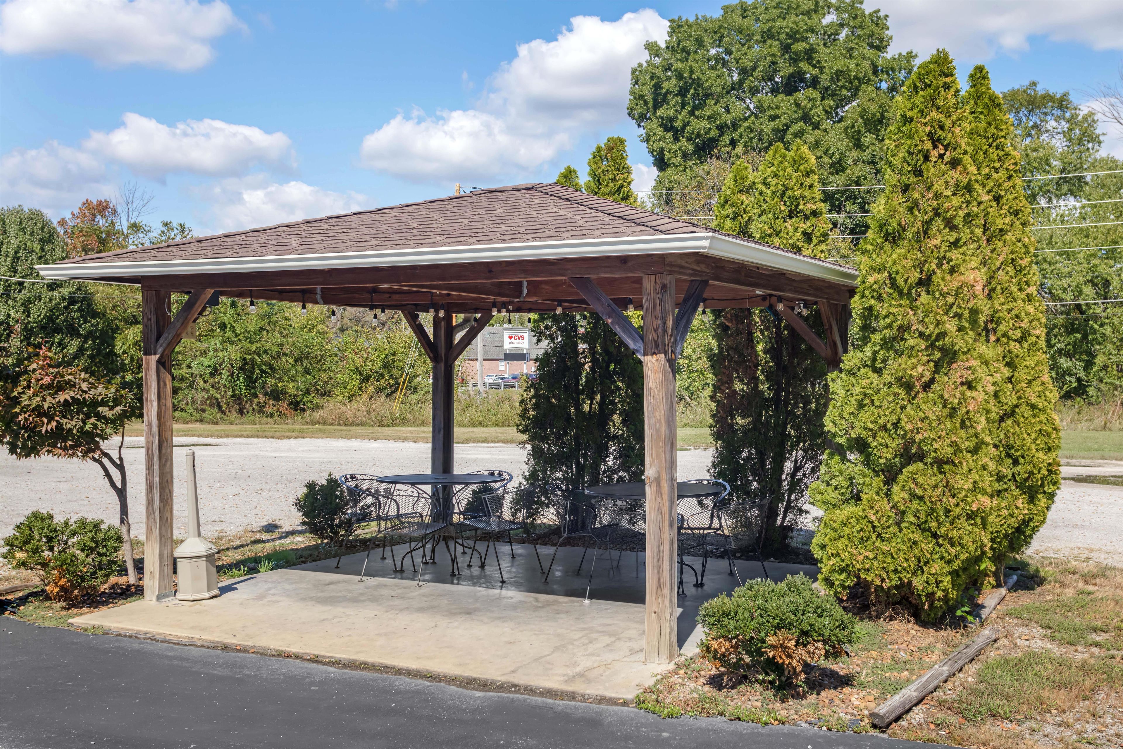 A gazebo with a tiled roof, surrounded by greenery and a paved area with outdoor seating.