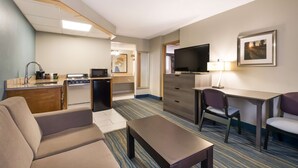 A compact hotel room with a kitchenette, a sofa, a desk with a chair, a television, and a framed picture on the wall.