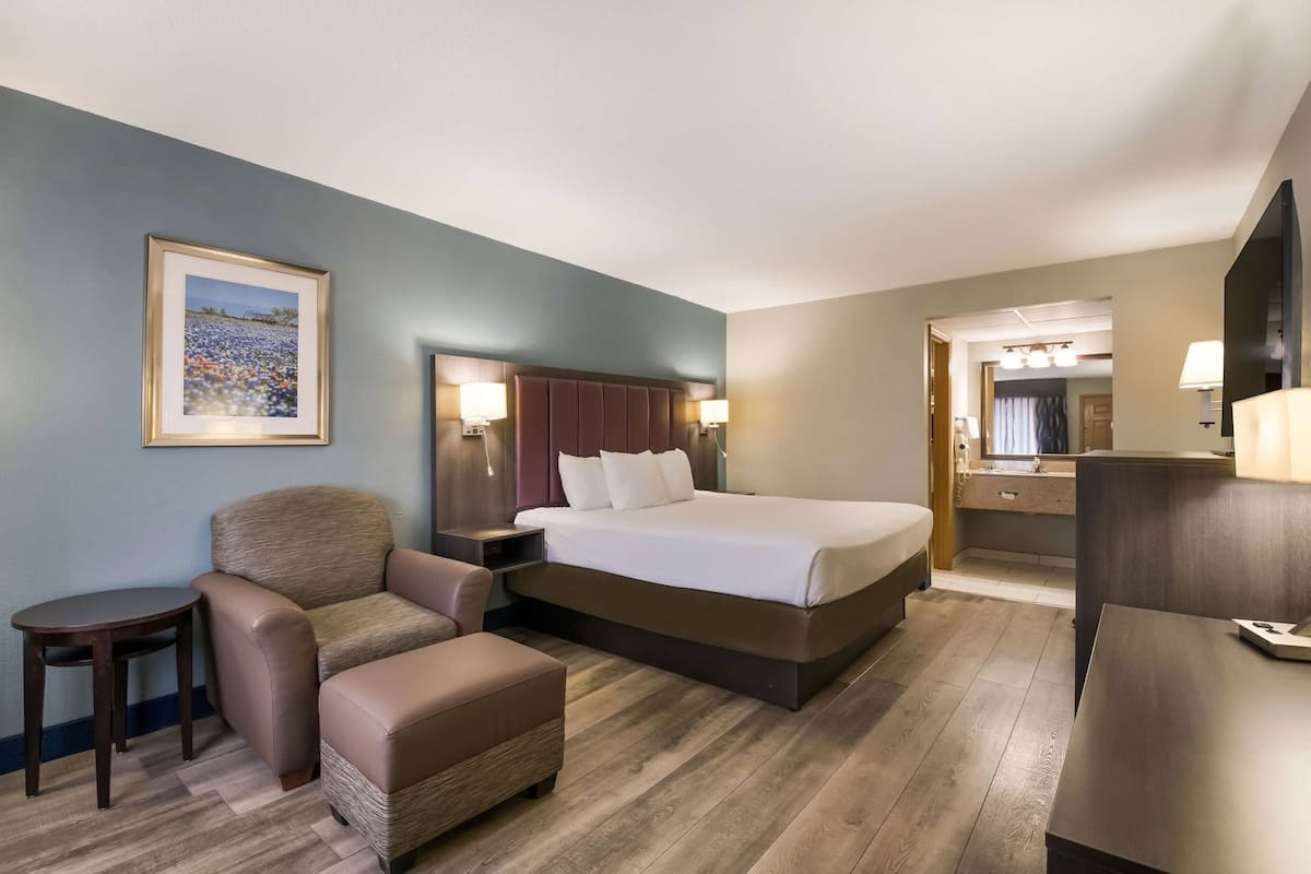 A hotel room with a bed, a chair, a small table, a nightstand, a lamp, a mirror, and a framed picture on the wall.