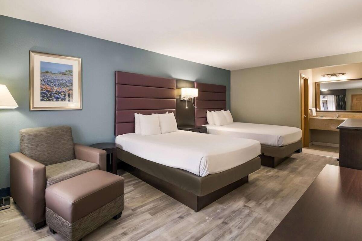 A hotel room with two beds, a chair, a desk, a lamp, and a framed picture on the wall.