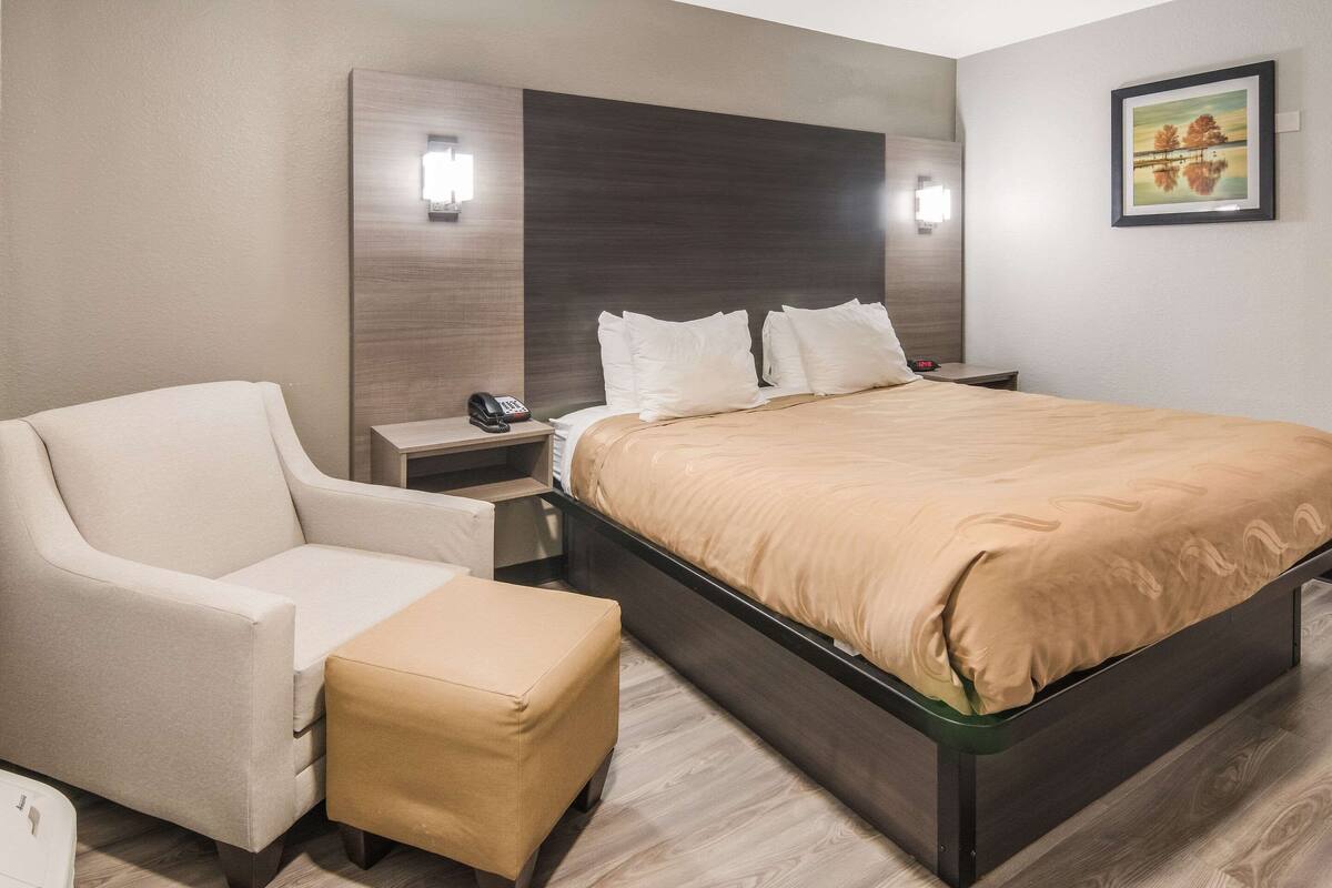 Ein modernes Hotelzimmer mit einem großen Bett, einem Nachttisch mit Telefon, einem Sessel und einem kleinen Hocker.