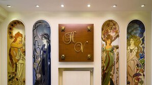 Un couloir orné de quatre panneaux de vitrail représentant des femmes vêtues à la mode classique, une plaque en bois portant les initiales « H. V. » et un radiateur.