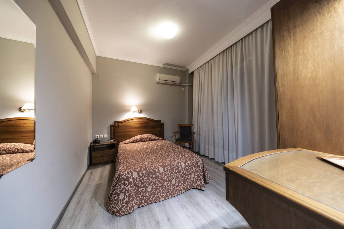 A hotel room with a wooden bed, a bedside table, a chair, and a large window with curtains.