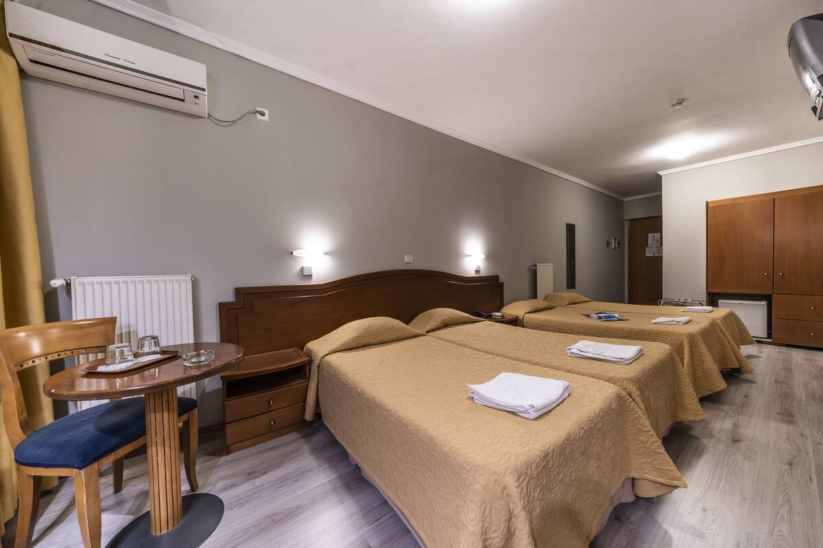 A hotel room with two beds, a wooden desk, a chair, a radiator, and a wall-mounted air conditioner.