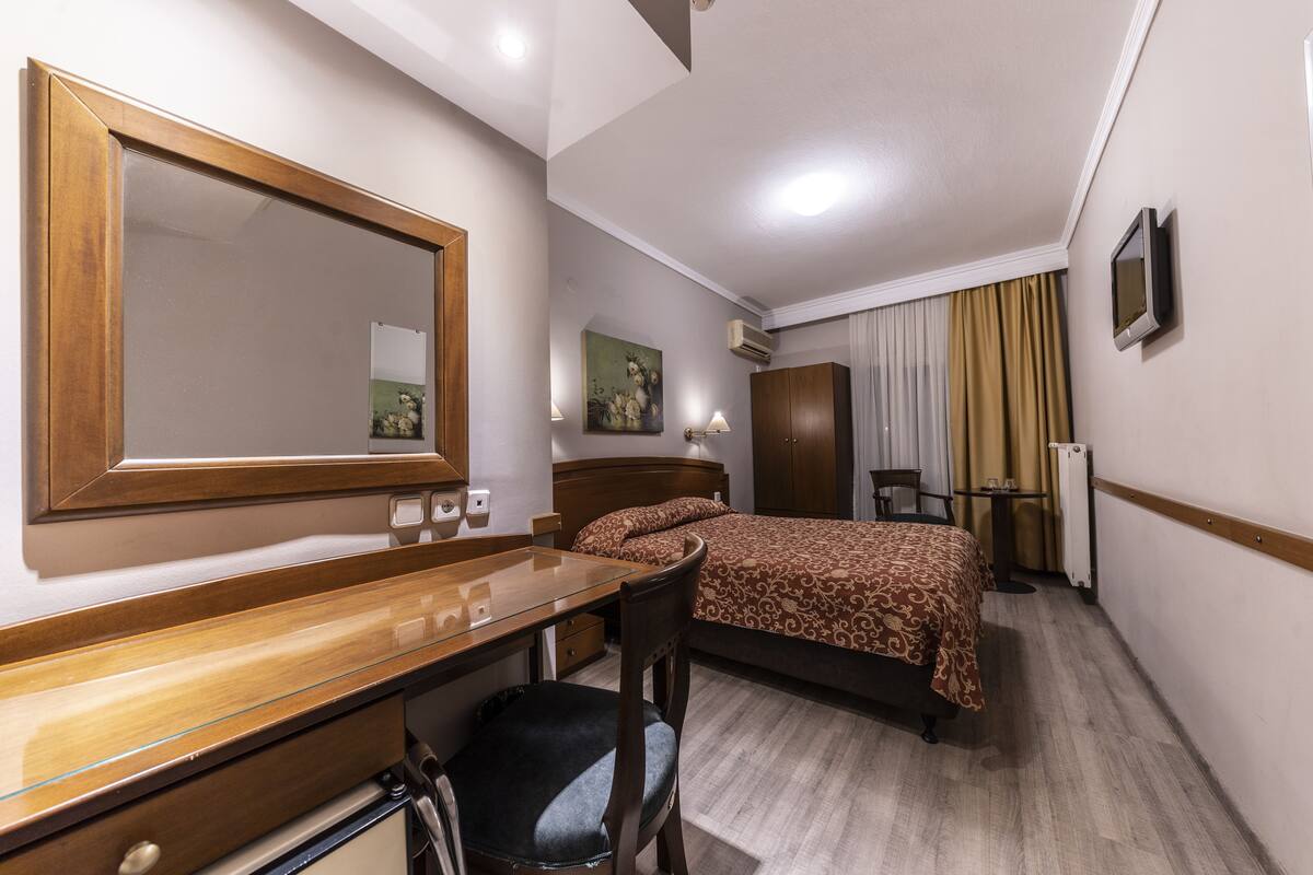 A hotel room with a wooden desk, a bed with a patterned bedspread, a chair, a mirror, a painting on the wall, and a window with curtains.