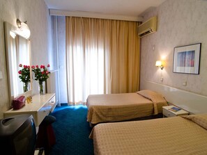 A hotel room with two beds, a desk with a mirror, a chair, a dresser, a trash bin, and a window with curtains. - Vergina Hotel (Thessaloniki)