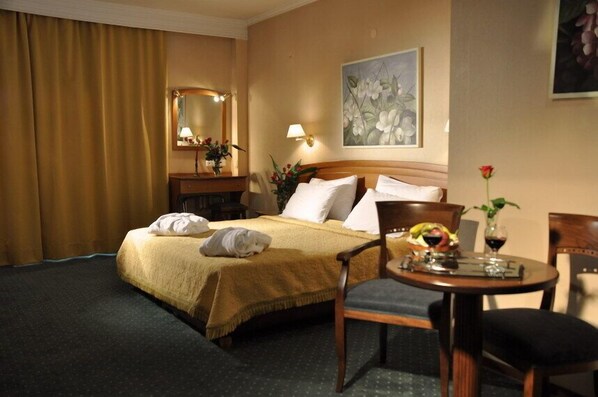 A hotel room with a bed, a round table with a vase and a fruit bowl, a chair, a mirror, and a painting on the wall. - Vergina Hotel (Thessaloniki)