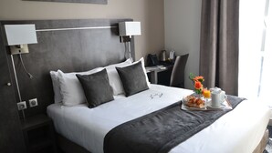 Una cama bien hecha con edredón blanco y gris, dos lámparas de noche, una mesita con una bandeja para el desayuno y una silla.