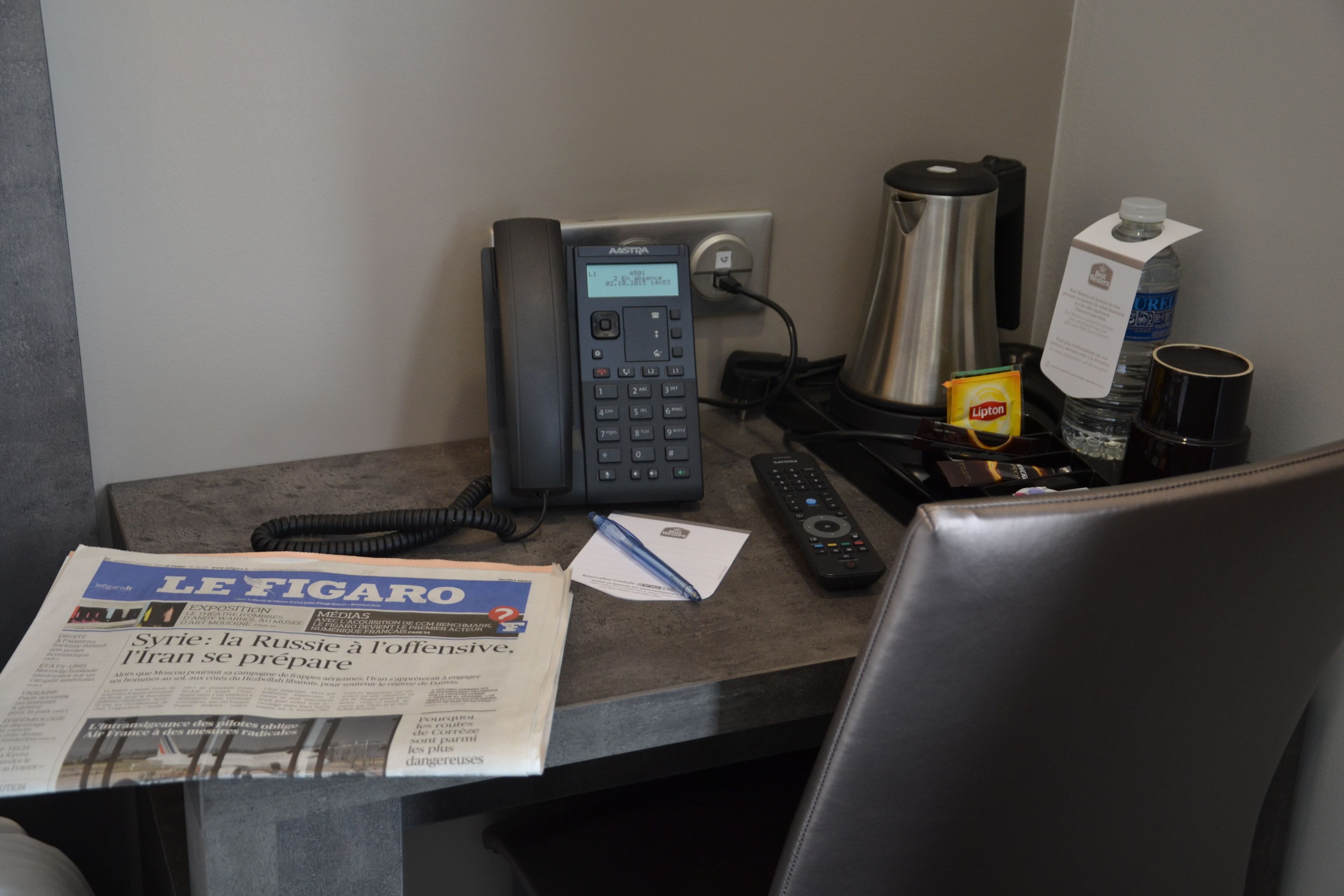 Escritorio de hotel con teléfono fijo, control remoto, periódico, cafetera y paquete de té Lipton.