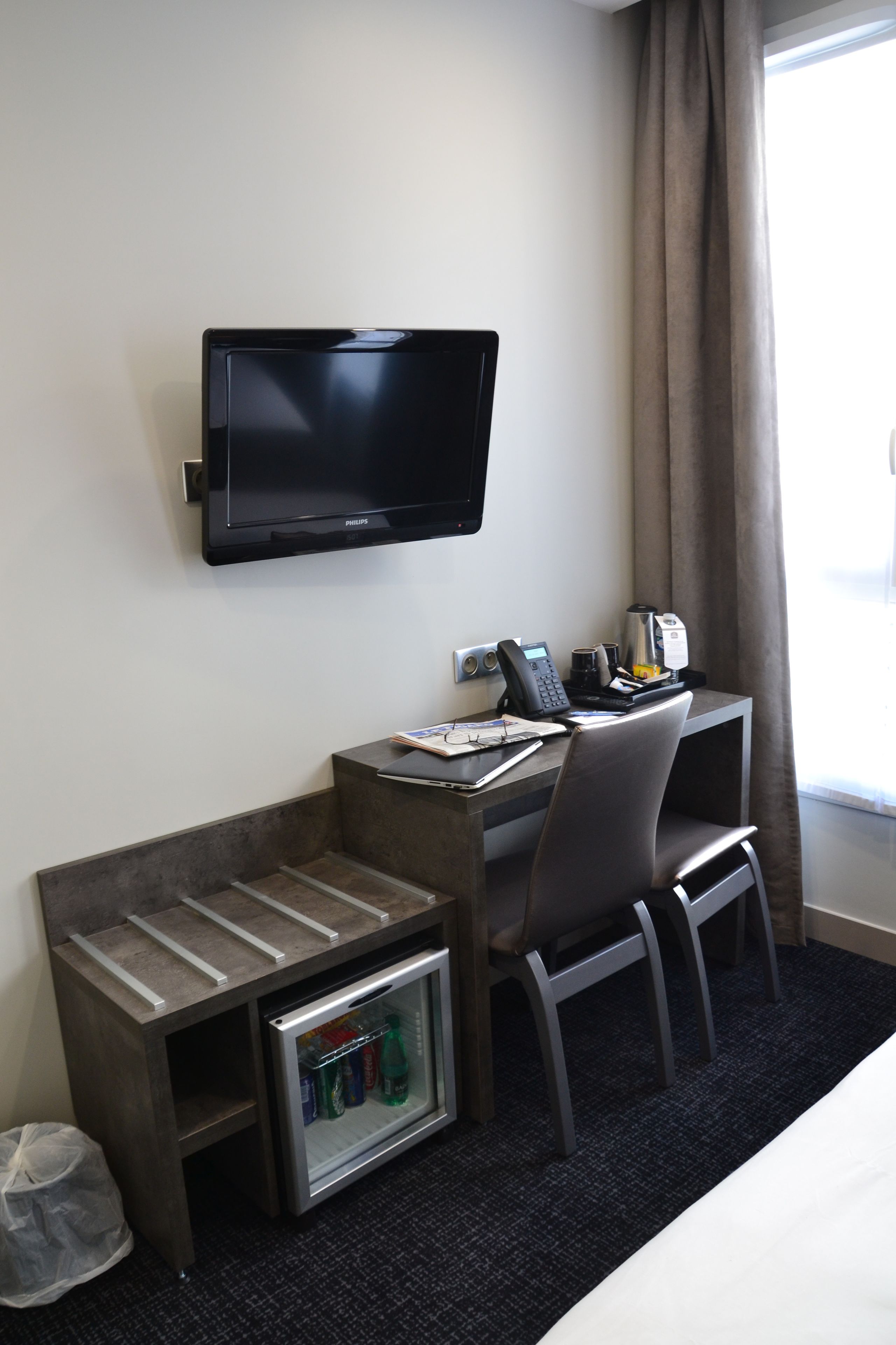 A hotel room with a desk, chair, television, mini-fridge, and a trash bin.