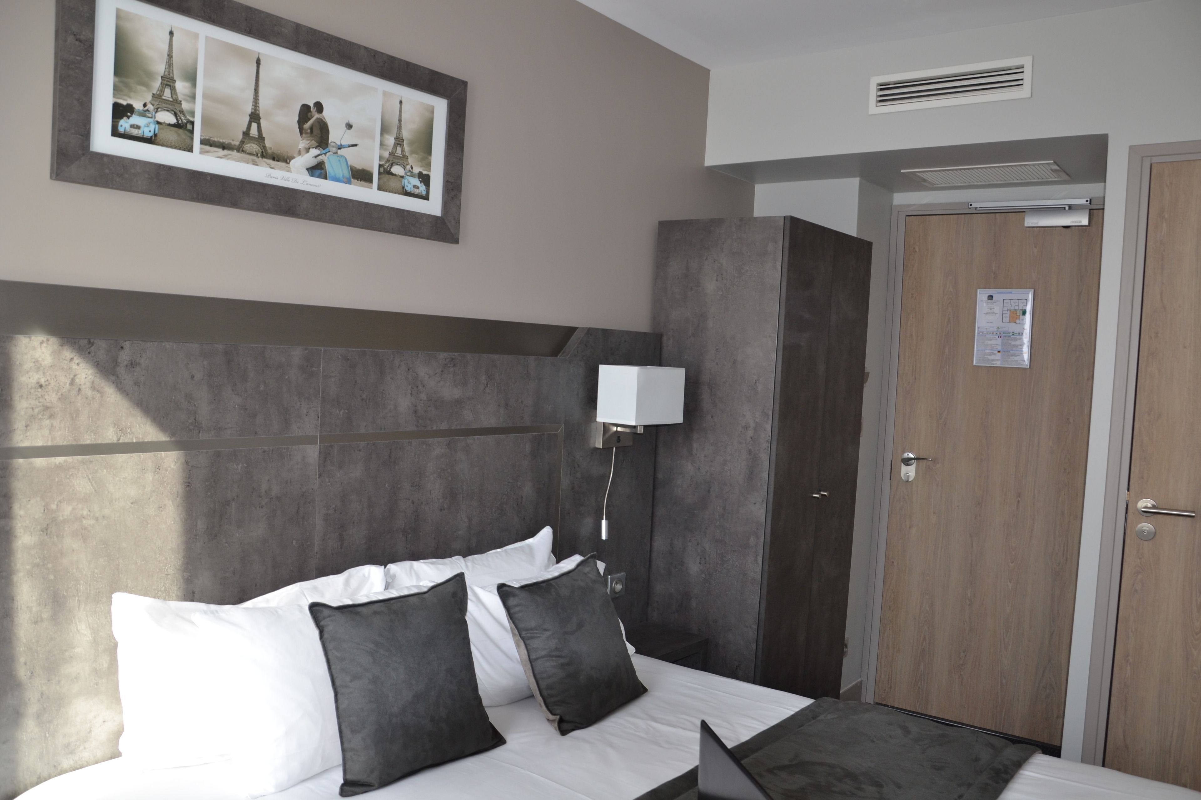 A hotel room with a bed, a nightstand, a wall-mounted lamp, a door, and a framed picture of the Eiffel Tower.